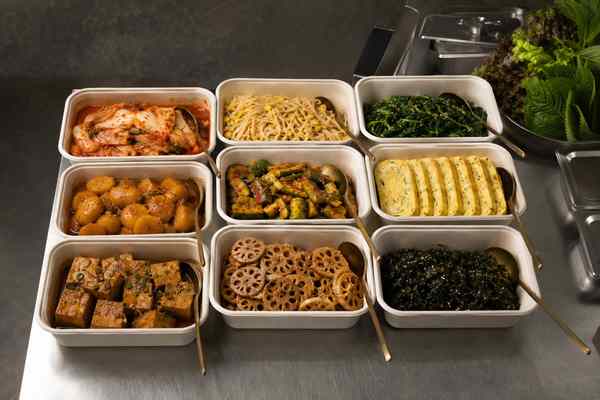 Nine open rectangular containers of assorted prepared banchan, each with a brass serving spoon, on a stainless counter; lettuce and perilla behind.