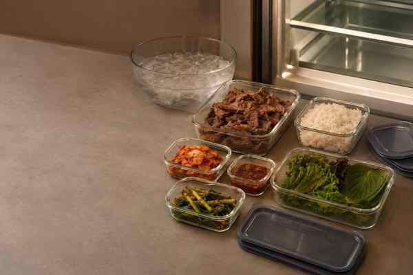 Open glass containers of bulgogi, rice, kimchi, seasoned greens, and lettuce on a counter, with separate lids, a bowl of ice, and an open refrigerator.