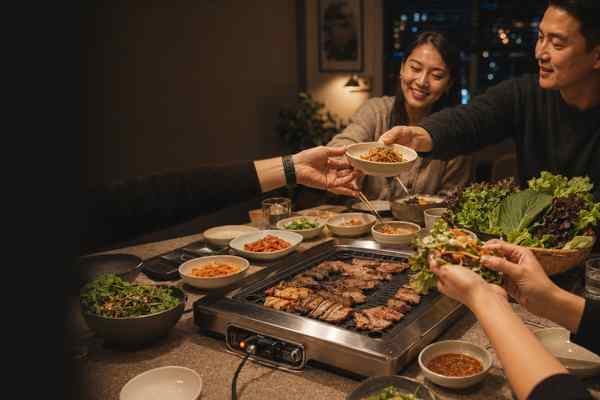 Adult friends passing lettuce wraps and banchan across a warmly lit Korean barbecue table.