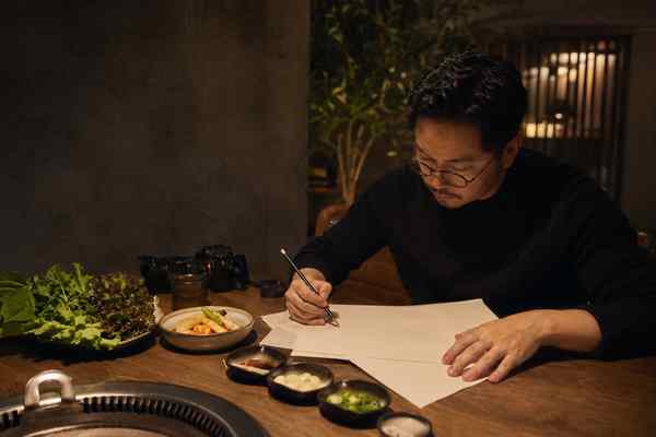 An adult editor writing on a blank cream page beside a warm Korean barbecue table.