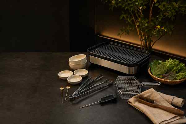 A curated Korean barbecue equipment still life with a tabletop electric grill, thermometer, tongs, and cream ceramics.