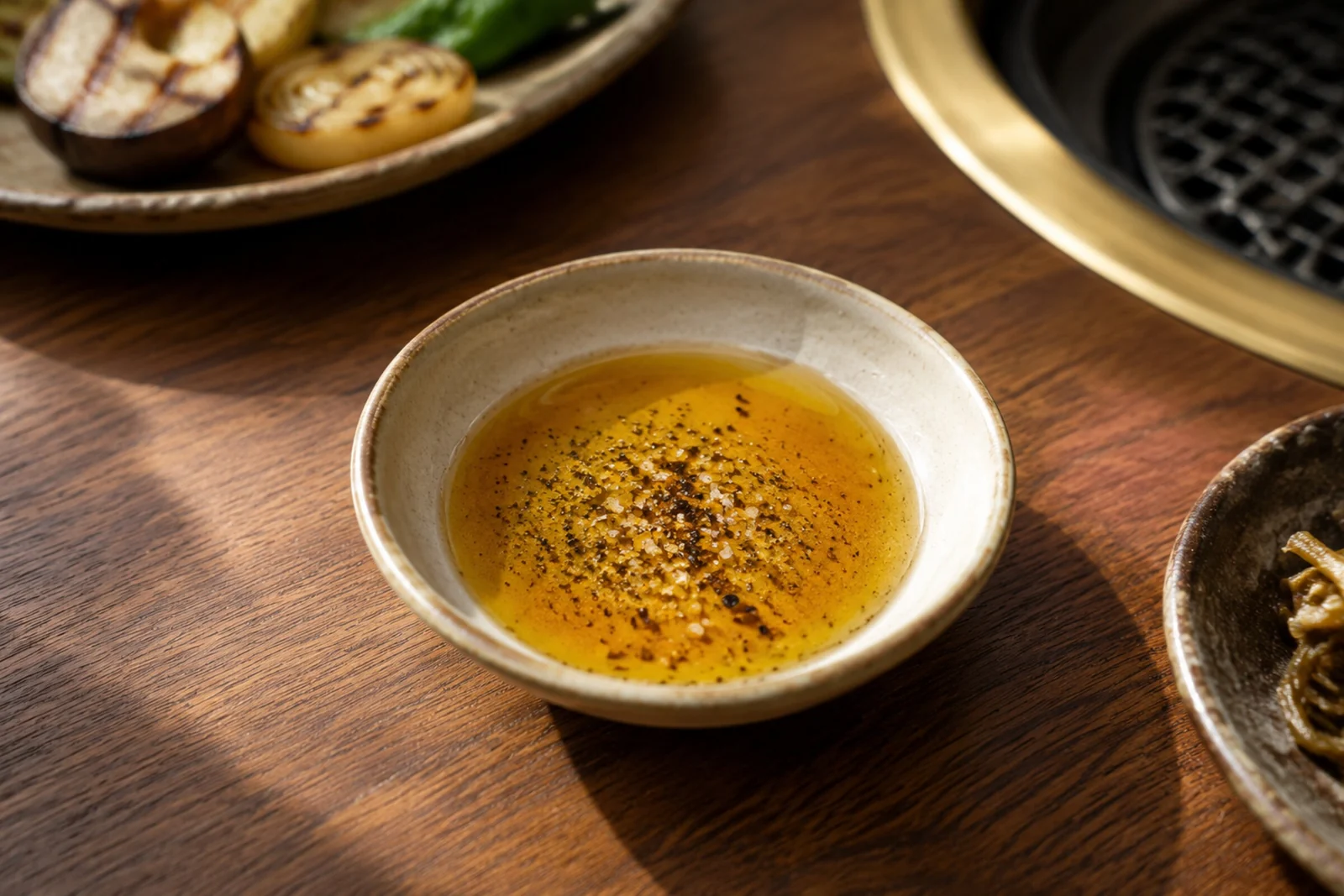 Small shallow dish of clear oil topped with salt and coarse black pepper, on a wooden table beside a plate of grilled vegetables and a grill.