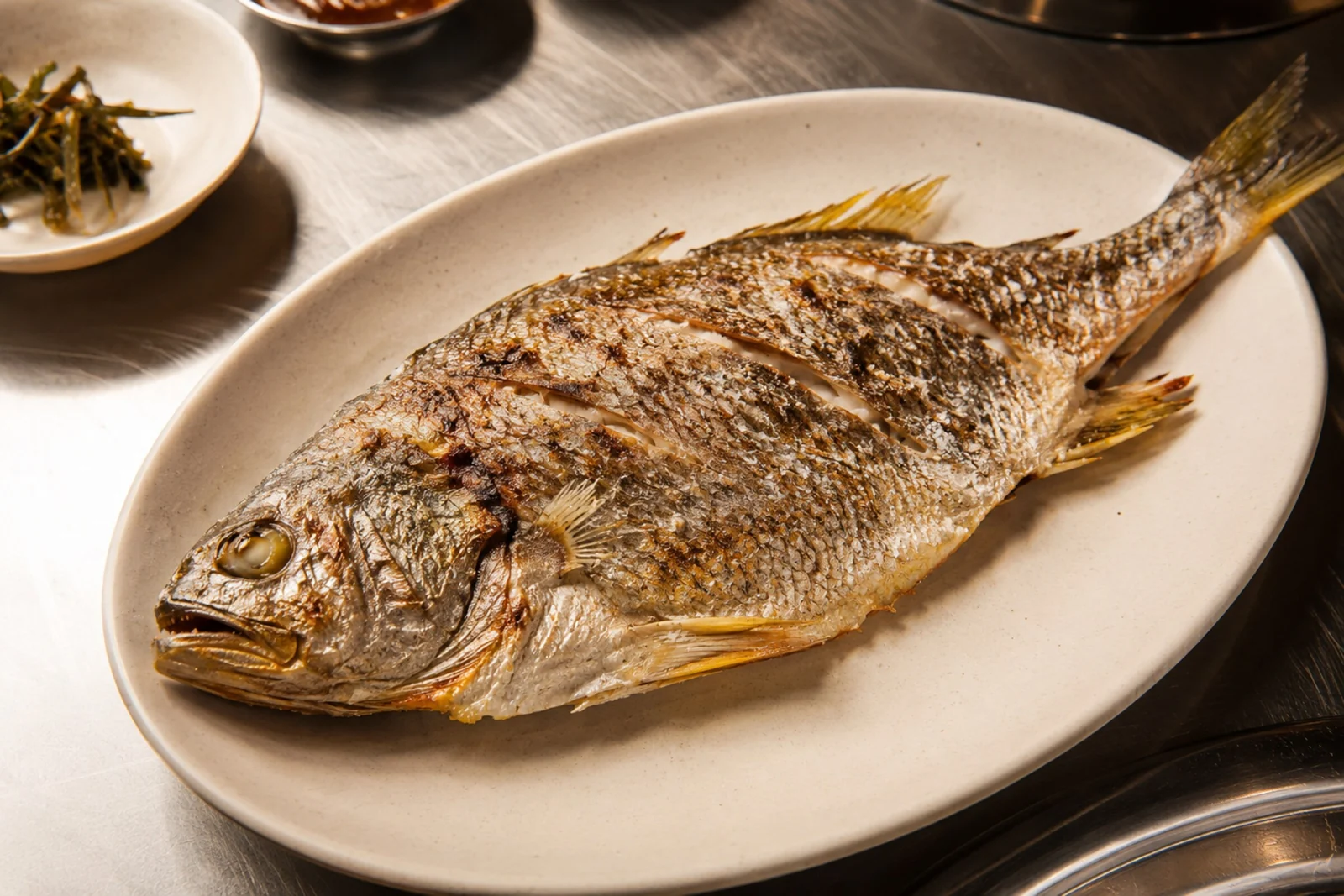 Whole grilled fish with crisp browned, scored skin on an oval plate, set on a stainless surface with a small side dish behind.